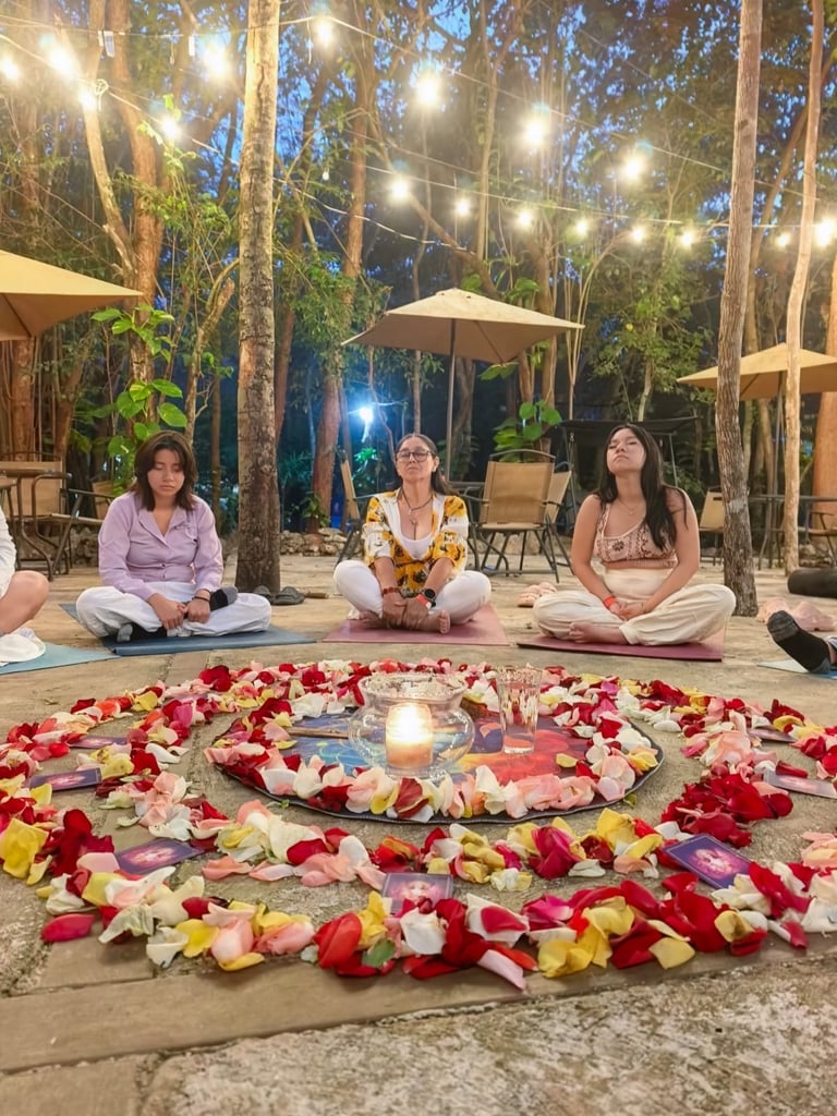 Three women sitting around a colorful flower petal mandala with candles at night in an outdoor garden setting with tall trees and string lights