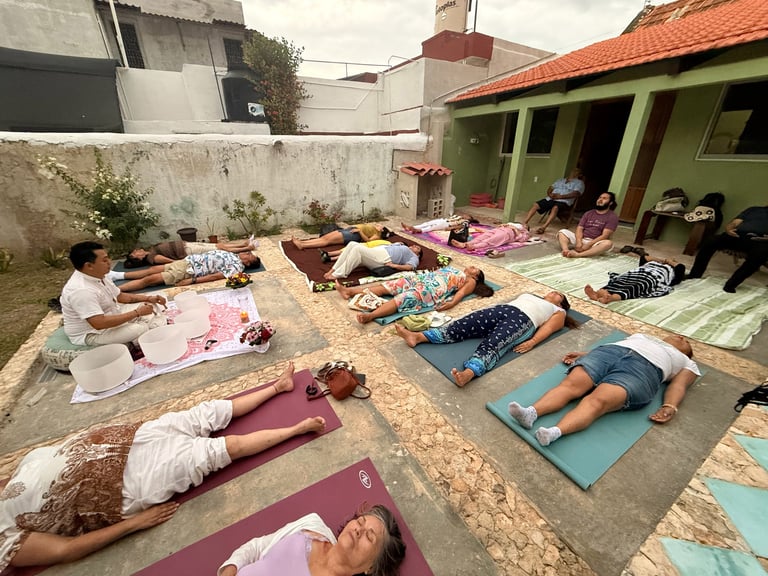 Group of people practicing yoga or meditation poses lying on mats in an outdoor courtyard with green buildings in background